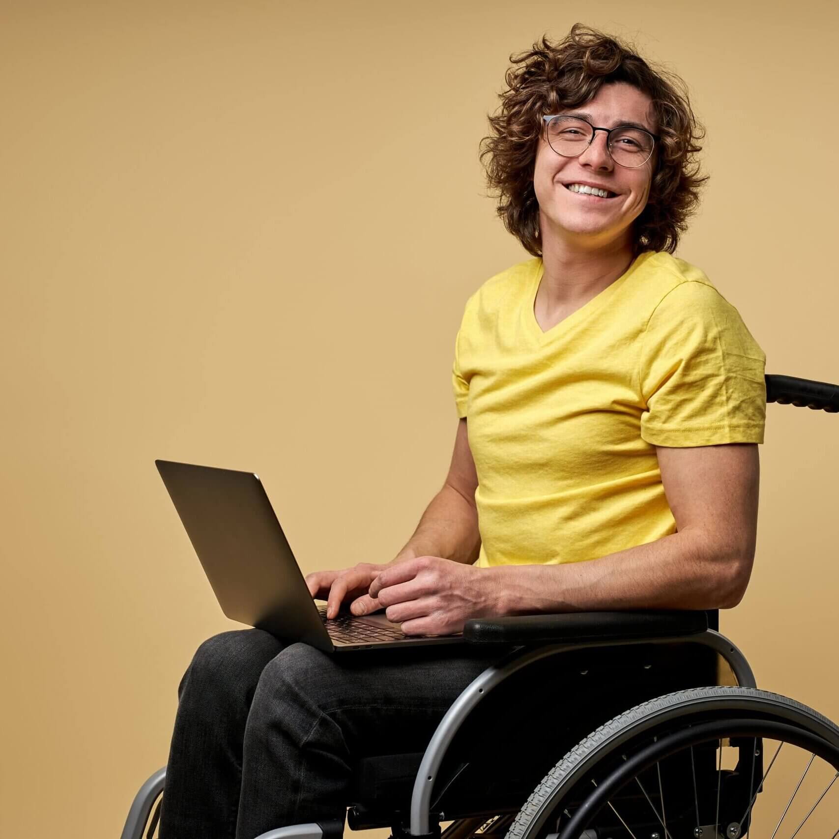 disabled man sits in a wheelchair with laptop, working online. curly young male in casual wear smiles at camera isolated in studio on beige background